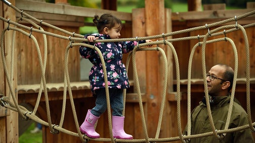 A young child in a winter coat walks along a rope bridge in the adventure play ground at Gibside while her dad watches on next to her.